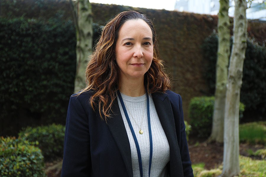Ana Quiñones, with long curly brown hair, a white top and black blazer, standing near greenery outside OHSU CHH1.