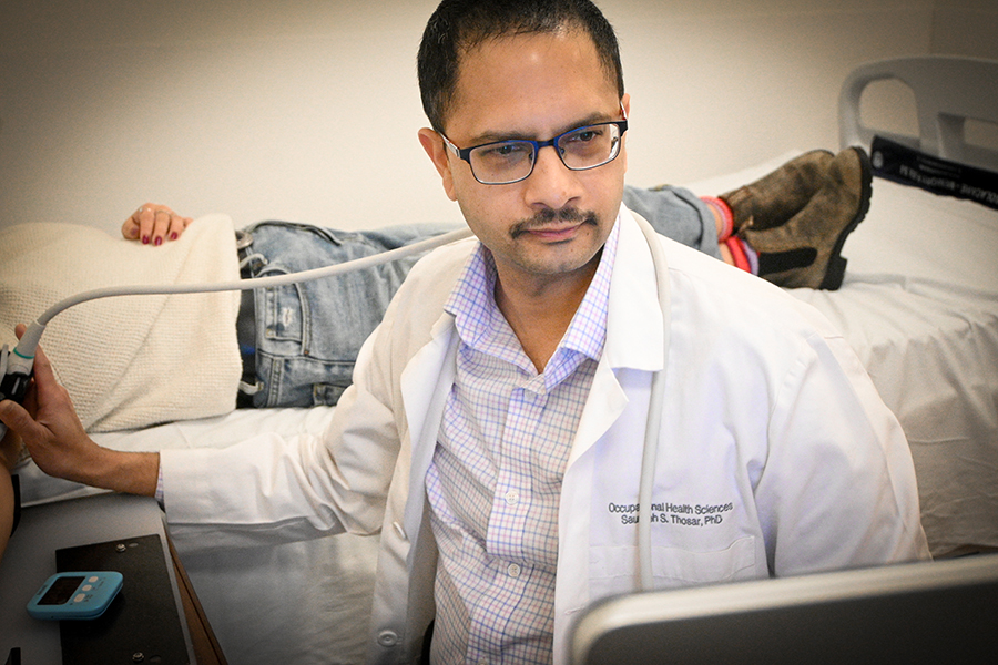 A man in a white lab coat sits bedside next to a patient lying down as he uses an instrument to collect sleep data.