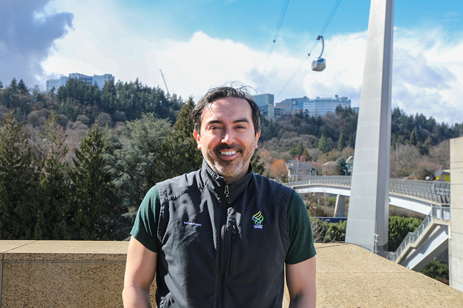 A man wearing an OHSU-branded vest smiles while standing outside with the OHSU Tram and Marquam Hill campus visible behind him.